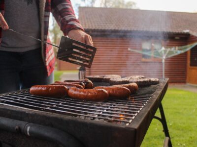 Grilling sausages and patties in a backyard with a rustic wooden house in the background.