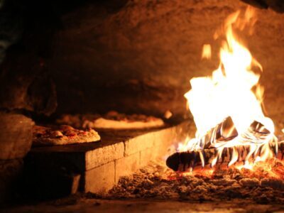 A close-up of a rustic wood-fired oven with pizzas near vibrant flames.