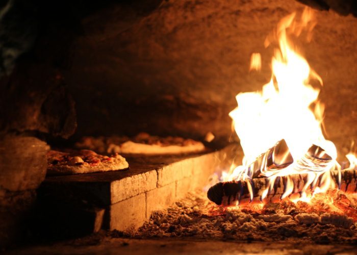 A close-up of a rustic wood-fired oven with pizzas near vibrant flames.