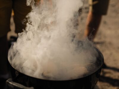 A smoky grill at an outdoor barbecue with people preparing food in a casual setting.