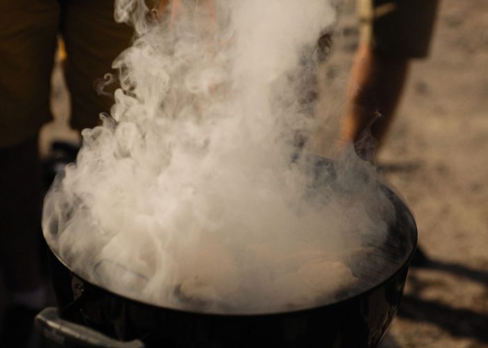 A smoky grill at an outdoor barbecue with people preparing food in a casual setting.